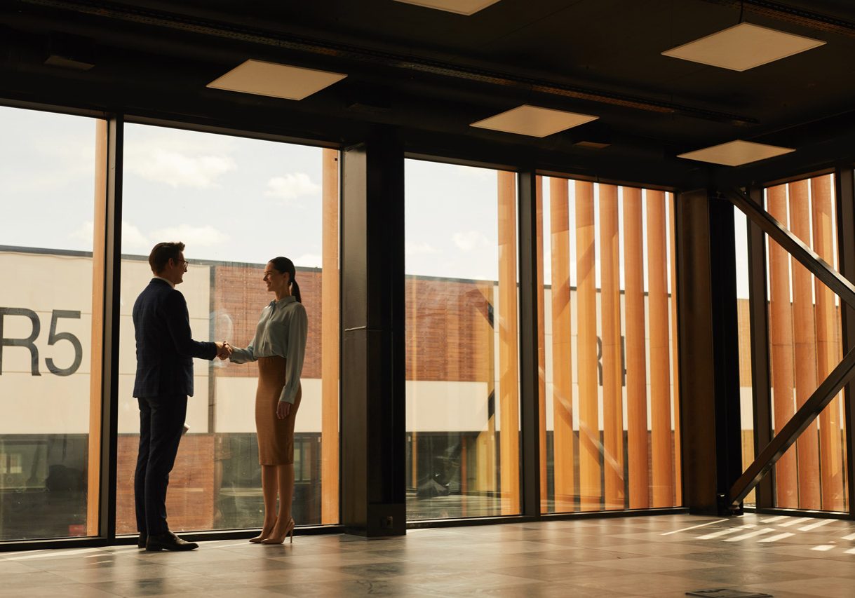 Wide angle view of real estate agent shaking hands with client while standing in empty office building interior lit by sunlight, copy space