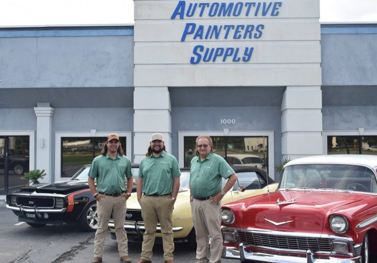 Three men in matching green shirts standing in front of classic cars outside the APS Mobile headquarters