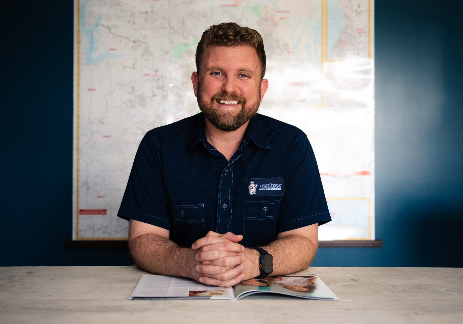 Josh Hembree smiling at desk in Hembree Heating & Air Conditioning uniform, in front of a large map
